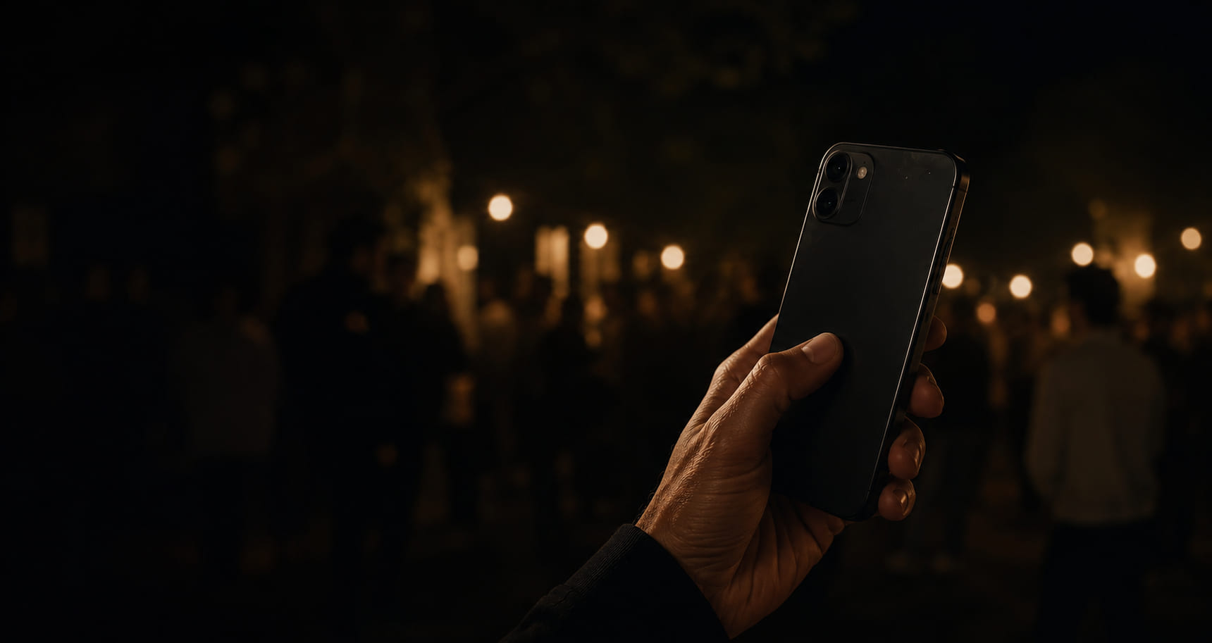 A hand holding a phone up at night in front of a blurred crowd, strings of warm bulbs behind them